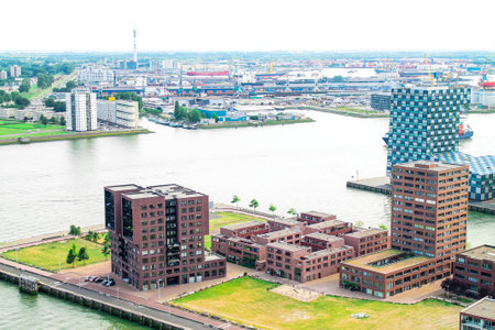 Aerial view of modern buildings along a waterfront, showcasing a mix of residential and commercial architecture. The scene includes a harbor with ships and industrial areas in the background, under a cloudy sky.の写真素材