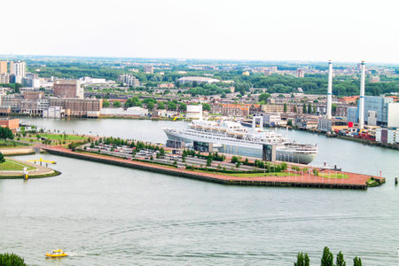 Aerial view of a harbor with a large cruise ship docked. The surrounding area features modern buildings, green spaces, and a river. In the background, industrial structures and a skyline are visible under a cloudy sky.の写真素材