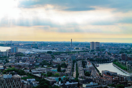 A panoramic view of a cityscape featuring a river, industrial buildings, and residential areas under a cloudy sky with rays of sunlight.の写真素材