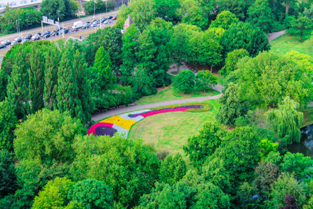 Aerial view of a lush green park featuring colorful flower beds arranged in patterns. The park is surrounded by dense trees and a winding path, with a glimpse of a road and cars in the background.の写真素材
