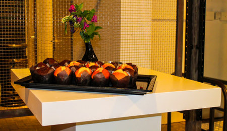 A modern table displaying a tray of assorted muffins, including chocolate and orange flavors, alongside a vase of flowers. The setting is stylish and contemporary, with a tiled background.の写真素材