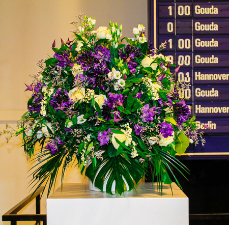 A vibrant floral arrangement featuring purple and white flowers, including roses and other blooms, displayed in a white vase. The background shows a board with information.の写真素材