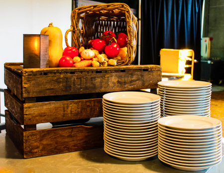 A rustic display featuring a wooden crate filled with fresh vegetables like tomatoes, squash, and mushrooms. Nearby, a stack of white plates is neatly arranged. The setting has a warm, inviting atmosphere.の写真素材