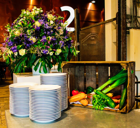A beautifully arranged display featuring a large floral arrangement in shades of purple and green, alongside stacks of white plates with blue rims. A rustic wooden crate holds fresh vegetables including cucumbers, carrots, and tomatoes, adding a touch of freshness to the setting.の写真素材