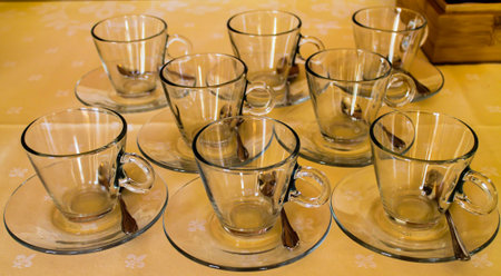 A set of eight clear glass cups with saucers arranged neatly on a light-colored tablecloth. Each cup has a small spoon placed on its saucer, creating a clean and elegant presentation.の写真素材
