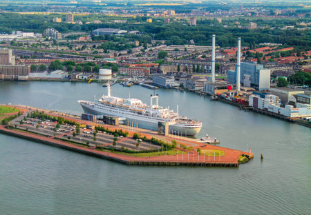 Aerial view of a large cruise ship docked at a harbor, surrounded by a cityscape with green areas and industrial buildings in the background. The water reflects the sky, creating a serene atmosphere.の写真素材