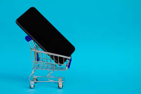 Shopping cart with black mobile phone on blue background. Copy space