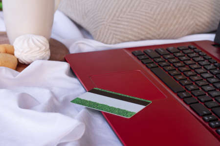Credit card on the keyboard of a red laptop standing on the bed next to a cup, marshmallows and cookies. Online shopping conceptの写真素材
