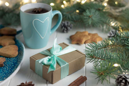 Gift wrapped in craft paper with mint ribbon, cup of tea with heart, gingerbread, anise, cinnamon next to green branches of a Christmas tree with LED light bulbs garland on a white wooden background. Merry Christmas and Happy New Year conceptの写真素材