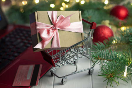 Shopping trolley with a gift near the keyboard of a red laptop, credit card, green branches of a Christmas tree with LED light bulbs, garland and shiny balls on a white wooden background. Merry Christmas and Happy New Year online shopping conceptの写真素材