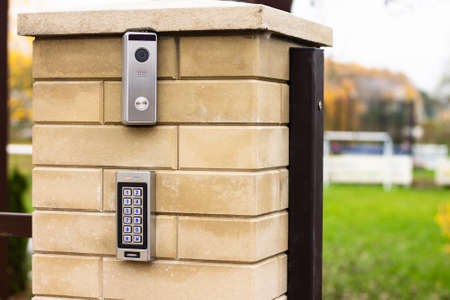 Silver intercom call panel with blue number buttons and a video camera on a brick beige fence pillar...