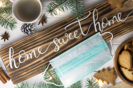 Home Sweet Home lettering wood sign with gingerbread cookie, Christmas tree branches, cup of coffee, cinnamon sticks and medical mask on white surface. Flat lay. Quarantine during a pandemic conceptの写真素材