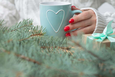 A woman's hand in a white knitted sweater with red nail polish holds a mint mug with a heart, near the branches of a New Year tree and a gift.の写真素材