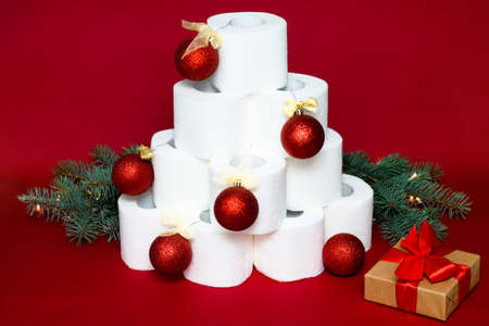 Christmas tree made from rolls of toilet paper, decorated with shiny balls, near a gift and fir branches on a red backgroundの写真素材