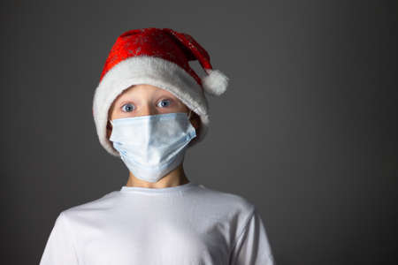 A 7-10 boy in a white T-shirt, Christmas cap and medical mask on a gray background. Christmas and new year during coronavirus pandemic concept. Copy spaceの写真素材