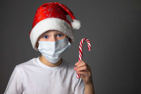 A 7-10 boy in a white T-shirt, Christmas cap and medical mask holding lollipop on a gray background. Christmas and new year during coronavirus pandemic concept. Copy spaceの写真素材