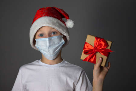 A 7-10 boy in a white T-shirt, Christmas cap and medical mask holding a gift on a gray background. Christmas and new year during coronavirus pandemic concept.の写真素材