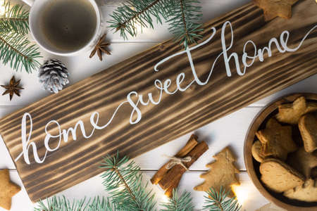 Home Sweet Home lettering burnt wood sign, gingerbread cookie, Christmas tree branches, cup of coffee, cinnamon sticks on white surface. Flat lay.の写真素材