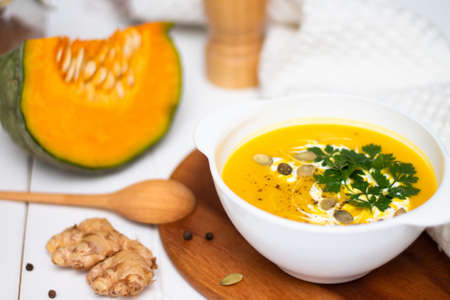 A plate of yellow pumpkin cream soup with cream, herbs and seeds on a white table near ginger, raw pumpkin and a wooden spoon.の写真素材