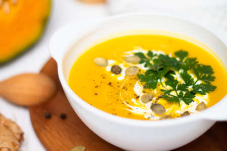A plate of yellow pumpkin cream soup with cream, herbs and seeds on a white table near ginger, raw pumpkin and a wooden spoon.の写真素材