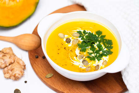 A plate of yellow pumpkin cream soup with cream, herbs and seeds on a white table near ginger, raw pumpkin and a wooden spoon.の写真素材