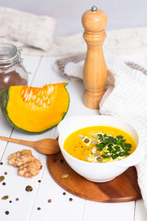 A plate of yellow pumpkin cream soup with cream, herbs and seeds on a white table near ginger, raw pumpkin and a wooden spoon. Verticalの写真素材