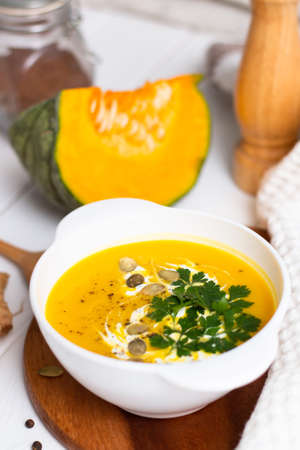 A plate of yellow pumpkin cream soup with cream, herbs and seeds on a white table near ginger, raw pumpkin and a wooden spoon. Verticalの写真素材