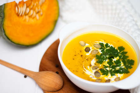 A plate of yellow pumpkin cream soup with cream, herbs and seeds on a white table near ginger, raw pumpkin and a wooden spoon.の写真素材