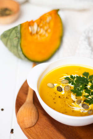 A plate of yellow pumpkin cream soup with cream, herbs and seeds on a white table near ginger, raw pumpkin and a wooden spoon. Verticalの写真素材