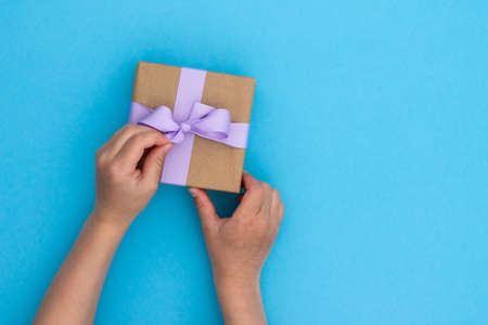 Hands receiving a gift in craft paper with a lilac ribbon on blue background. Top view. Copy spaceの写真素材