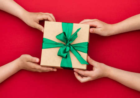 Hands giving or receiving a gift in craft paper with a green ribbon on red background. Top view. Copy spaceの写真素材