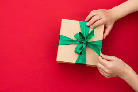 Hands giving or receiving a gift in craft paper with a green ribbon on red background. Top view. Copy spaceの写真素材