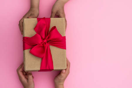 Hands giving or receiving a gift in craft paper with a pink ribbon on pastel pink background. Top view. Copy spaceの写真素材