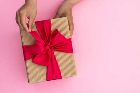 Hands giving or receiving a gift in craft paper with a pink ribbon on pastel pink background. Top view. Copy spaceの写真素材