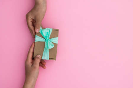 Hands giving or receiving a gift in craft paper with a mint ribbon on pastel pink background. Top view. Copy spaceの写真素材