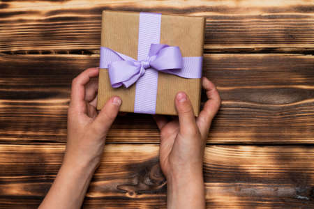Hands giving or receiving a gift in craft paper with a lilac ribbon on a rustic burnt wood background. Top view. Copy spaceの写真素材