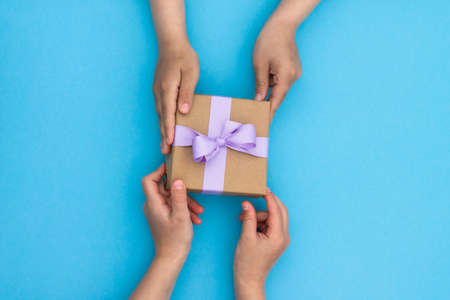 Hands giving or receiving a gift in craft paper with a lilac ribbon on blue background. Top view. Copy spaceの写真素材
