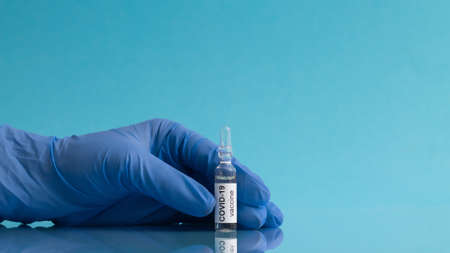 Hand in a medical glove holding an ampoule with the COVID-19 coronavirus vaccine on a blue background, copy space. Bannerの写真素材