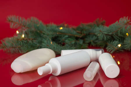 White bottles of cosmetics near christmas tree branch with lights on red surfaceの写真素材