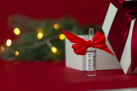 An ampoule of the COVID-19 coronavirus vaccine stands as a gift near a Christmas tree branch with lights on a red backgroundの写真素材