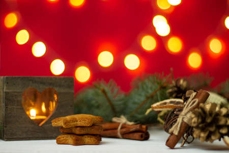 Heart shaped wooden candlestick with Christmas tree branches, gingerbread and cinnamon on white table and bokeh on a red background. Merry christmas and a happy new year. Copy spaceの写真素材