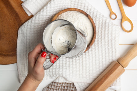 A woman's hand sifts the flour with a special mug with a sieve near a white towel, a wooden rolling pin and spoons. Flat layの写真素材