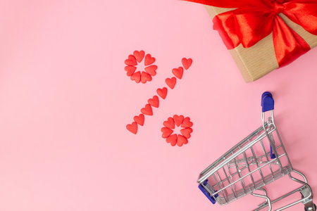 Percent sign lined with small red hearts with gift wrapped in craft paper and red ribbon and shopping cart on pink background. Flat lay. Copy space.の写真素材