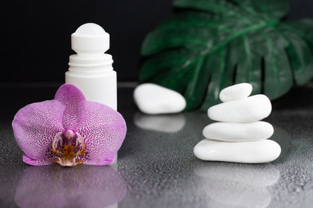 Beautiful lilac orchid flower and white roll-on deodorant with white stones and monstera leaves, on black wet backgroundの写真素材