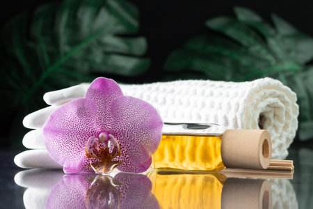 Beautiful lilac orchid flower, clear bottle of yellow oil or perfume, wooden sticks and rolled towel with stack of white stones and monstera leaves, on black surface. SPA salon and procedures conceptの写真素材