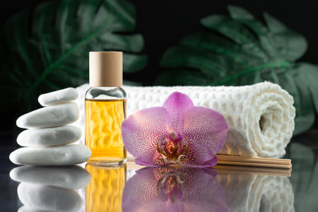 Beautiful lilac orchid flower, clear bottle of yellow oil or perfume, wooden sticks and rolled towel with stack of white stones and monstera leaves, on black surface. SPA salon and procedures conceptの写真素材