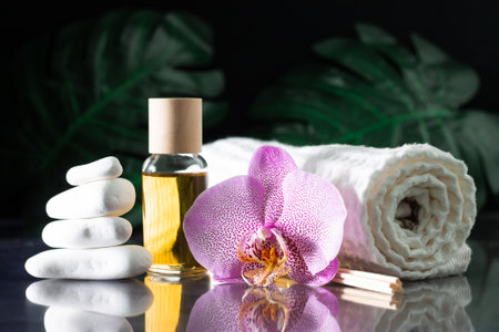 Beautiful lilac orchid flower, clear bottle of yellow oil or perfume, wooden sticks and rolled towel with stack of white stones and monstera leaves, on black surface. SPA salon and procedures conceptの写真素材