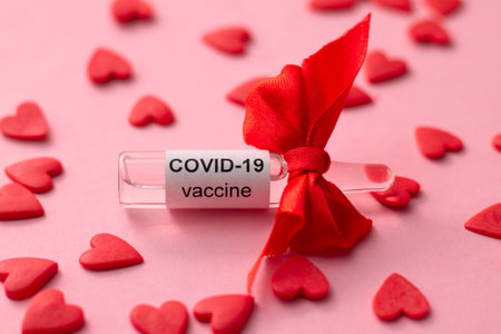 Ampoule with COVID-19 vaccine as gift for Valentine's Day next to red hearts on pink backgroundの写真素材