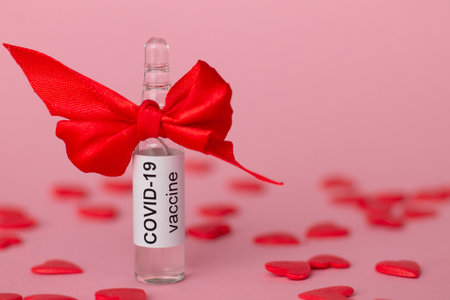 Ampoule with COVID-19 vaccine as gift for Valentine's Day next to red hearts on pink background. Copy spaceの写真素材