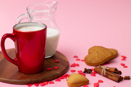 Red latte cup and jug of milk with heart shaped cookies, cinnamon, anise and red hearts on pink background. Copy spaceの写真素材